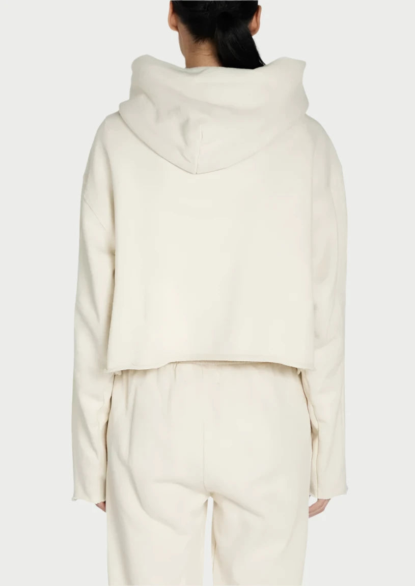 A person with dark hair tied back in a ponytail is facing away, wearing a cream-colored, hooded sweat top and matching bottoms. The 100% cotton Rosa Raw Zip Hoody by Les Tien appears to be a crop hoodie featuring a RIRI zipper, and the bottoms are high-waisted. The background is plain white.