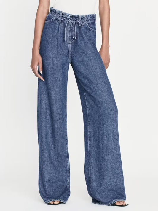 A person is wearing Frame's Super Drape Drawstring Pants, which feature a high-waisted, wide-leg design and a drawstring waist. The medium blue pants are made of lightweight denim, and the person stands against a plain white background. Their upper body is not fully visible in the image.