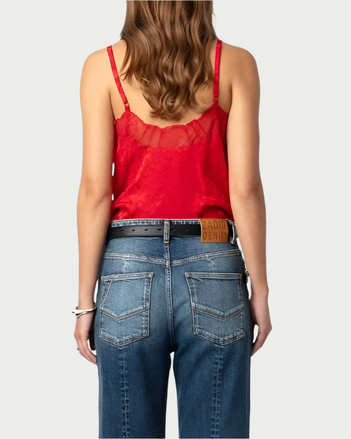 A woman with wavy hair, facing away from the camera, showcases a Zadig & Voltaire Crimson Cyoko Leopard Print Cami paired with blue jeans featuring a branded leather patch. She accessorizes with a black belt and bracelets on her left wrist, adding elegance to her look.