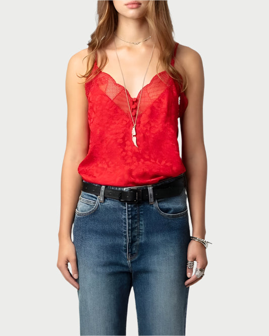 A person wearing the Crimson Cyoko Leopard Print Cami by Zadig & Voltaire and blue jeans stands against a plain background. Accessories include a black belt, silver bracelets, rings, and layered necklaces. The look is completed with loose hair for an effortless touch.