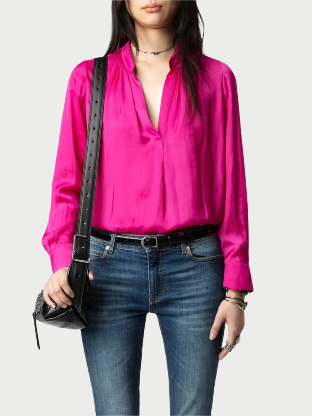 A person dressed in a vibrant Zadig & Voltaire Bright Pink Satin Blouse featuring a deep V-neck and long sleeves, complemented by blue jeans and adorned with bracelets, a choker necklace, as well as carrying a black leather bag.