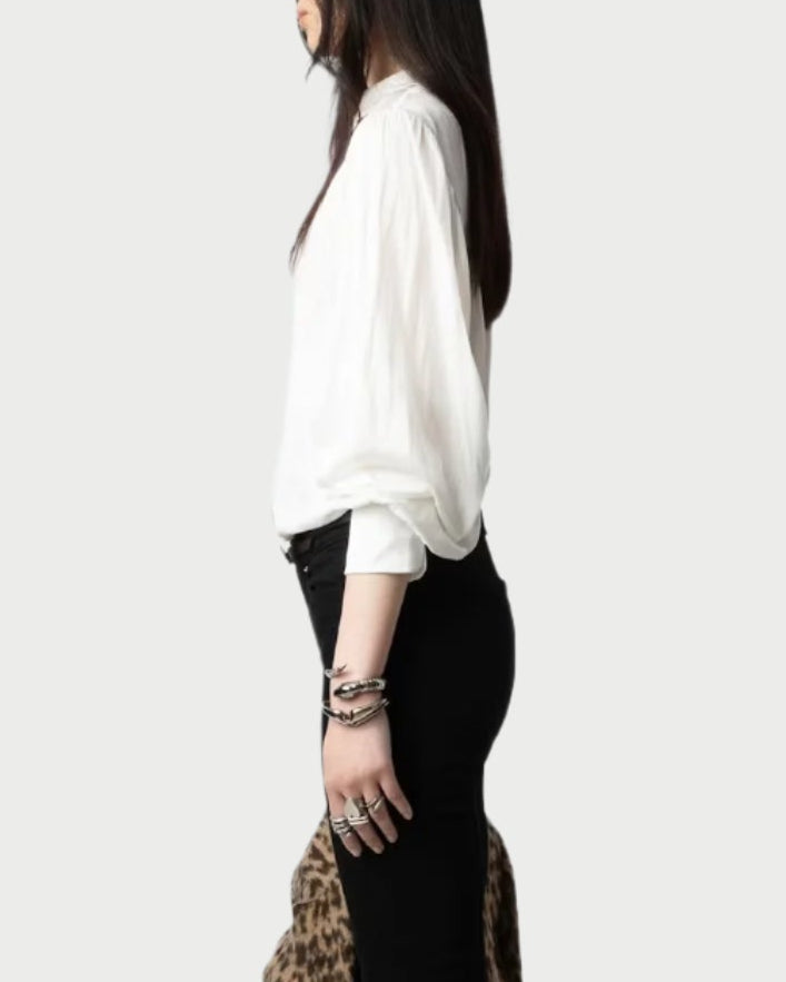 A person stands in profile, wearing a Trenta Satin Blouse by Zadig & Voltaire and black pants against a plain background. They have long dark hair, accessorized with multiple silver bracelets and rings. A recycled polyester leopard-print fabric is partially visible at the bottom.