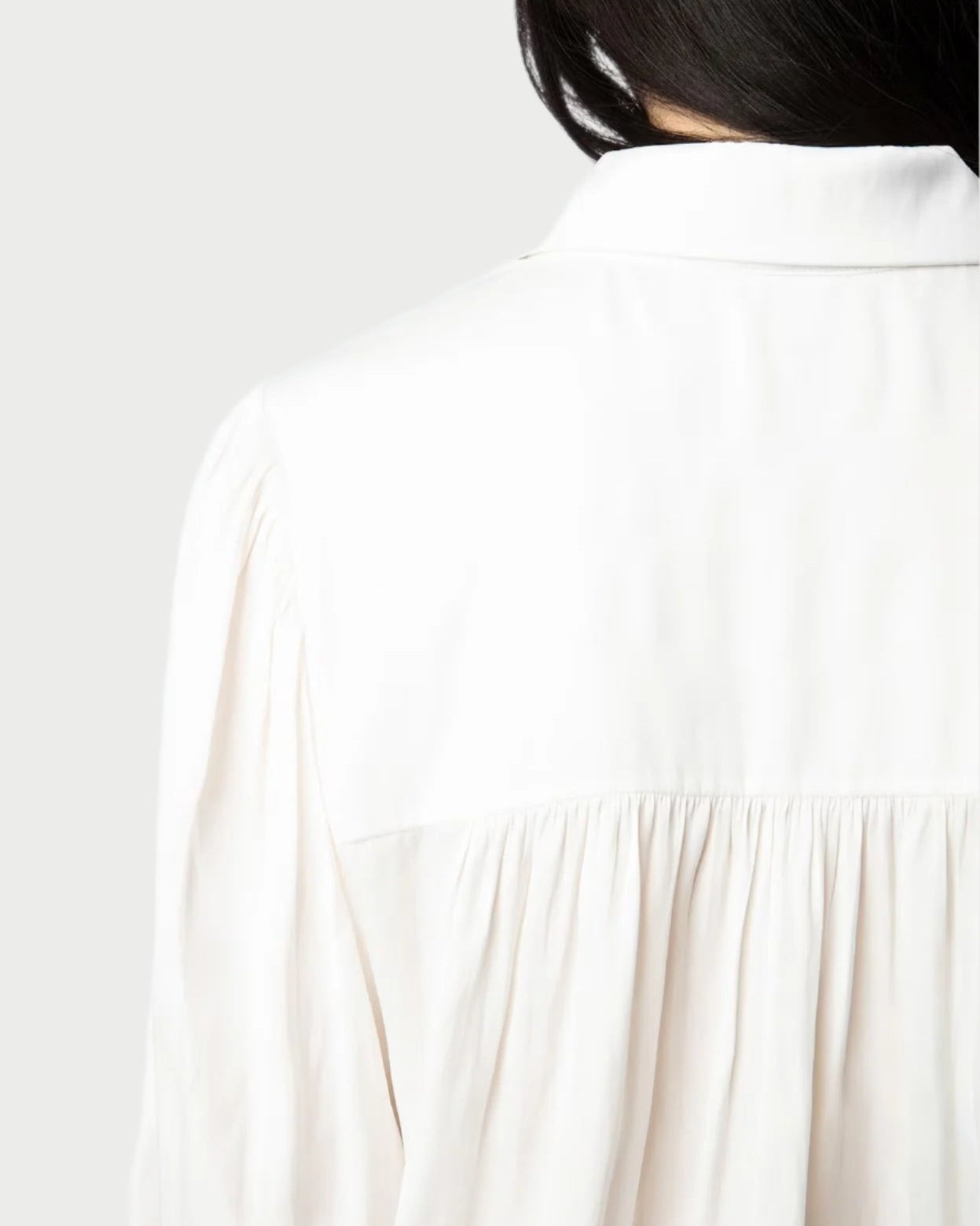 A close-up shows a person wearing the Zadig & Voltaire Trenta Satin Blouse, highlighting its gathered fabric from shoulder to mid-back against a muted background. The piece pairs elegance with sustainability, featuring long dark hair for contrast.