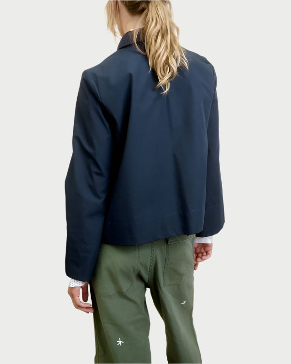 A person with long blonde hair wears an AqC Cropped Jacket over a white shirt and green pants. The boxy fit of the navy jacket enhances the ensemble against a light gray backdrop, all shown from behind.