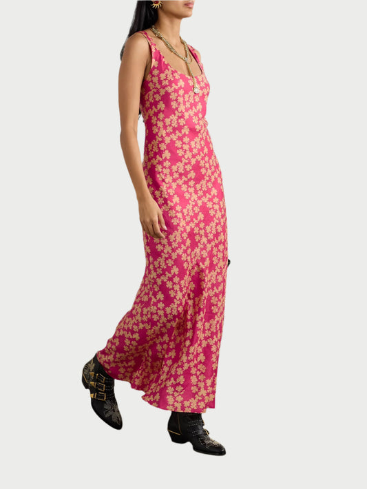 A woman elegantly models the Adele Dress by Alix of Bohemia, a sleeveless, long pink 100% silk bias-cut slip with white floral patterns. She pairs it with black ankle boots adorned with gold details. Her long black hair flows freely as she accessorizes with large earrings and layered necklaces.