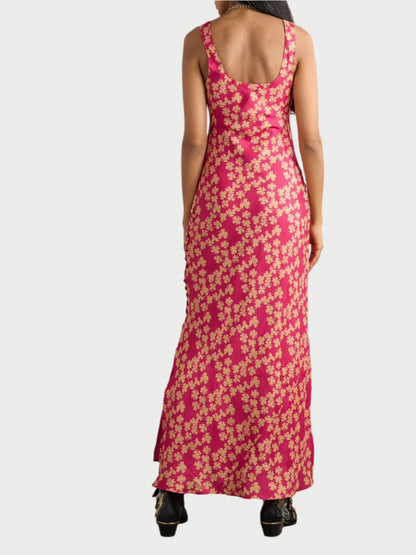 The person is seen from behind wearing the Adele Dress by Alix of Bohemia, a sleeveless pink maxi dress with a yellow floral pattern. It features a hand-block printed design, scoop neckline, and ankle-length flow, styled with black shoes against a plain background.