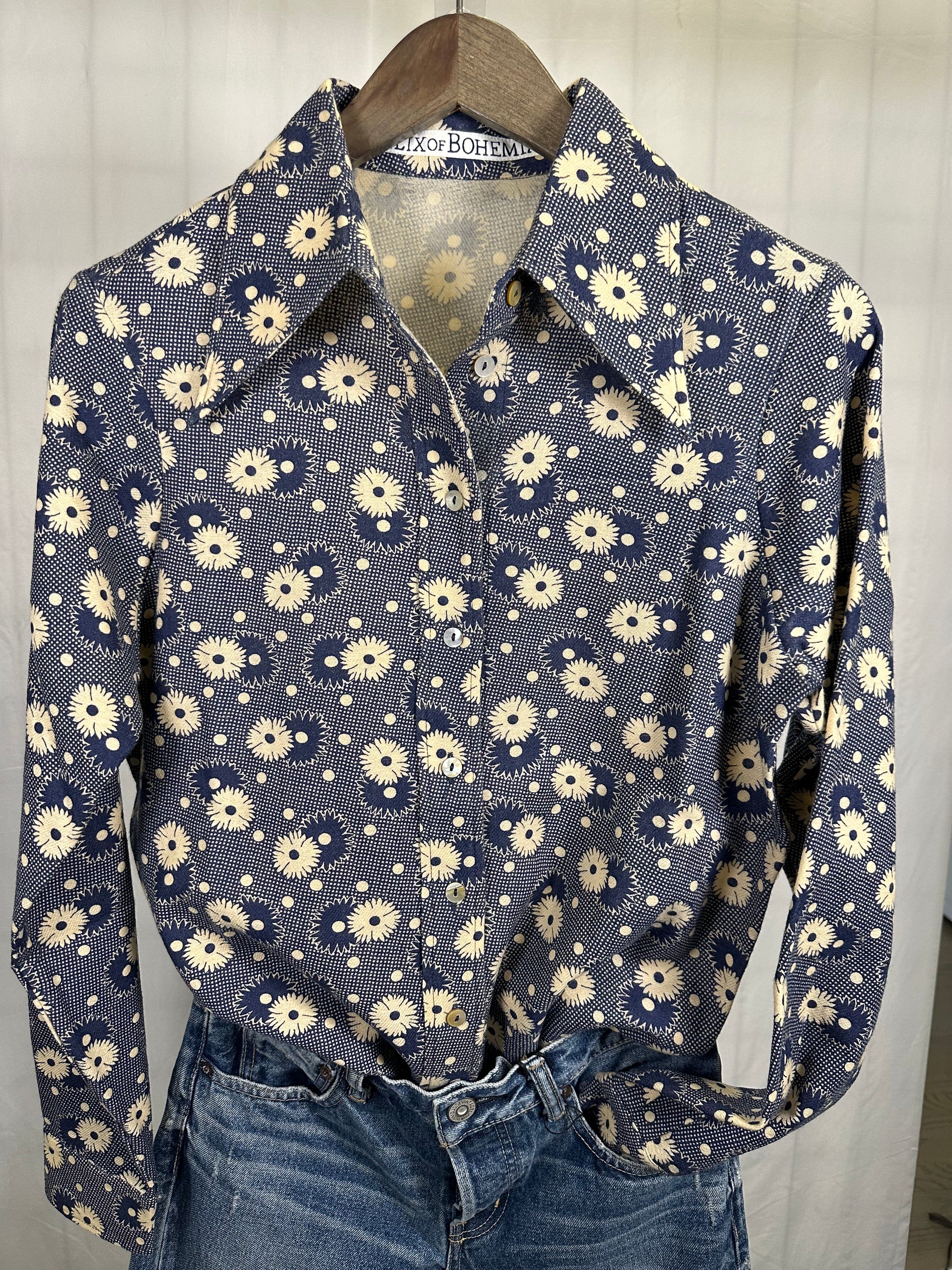 An Alix of Bohemia Daisy Print Shirt, featuring a navy blue background with long sleeves and adorned with daisy prints, hangs elegantly on a wooden hanger. Underneath the shirt, a pair of blue jeans completes the perfect fall button-down ensemble.