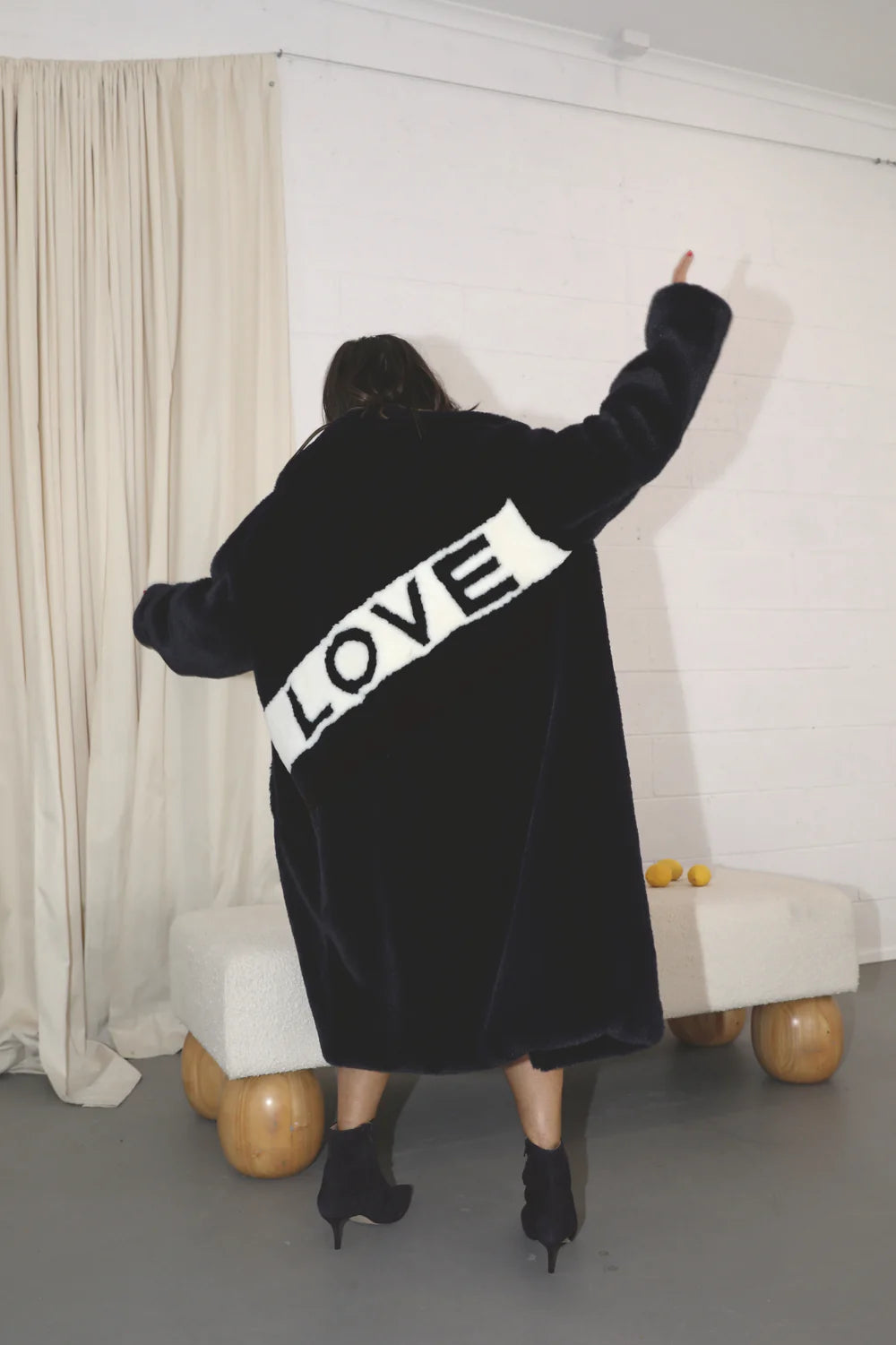 A person stands with their back to the camera, wearing a large black Mr Greys Love Coat from ANGEL WINGS, featuring the word "LOVE" in bold white letters across the back. The coat's faux fur collar adds a touch of elegance. The person is indoors, with a beige curtain and minimalist furniture in the background.