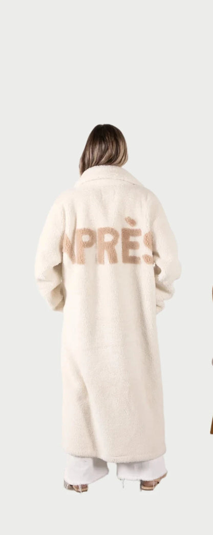 A person with long hair, clad in an Angel Wings Aprés Coat featuring the word "APRÈS" on the back, stands facing away from the camera. Beside them is a wooden chair with a woven backrest, all set against a plain white wall. The scene evokes a serene Winter Season ambiance.