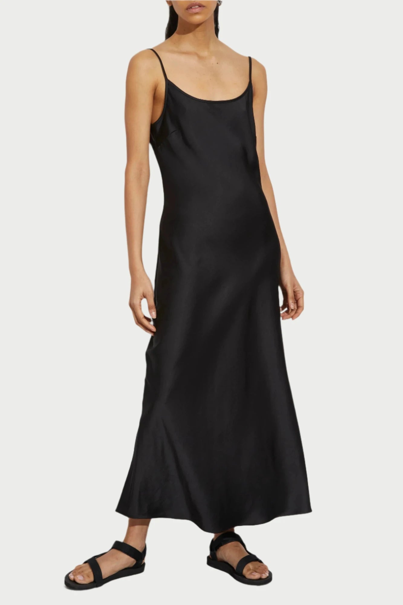 A person stands against a white background, wearing the Black Haley Slip Dress by Saint Art NY—a long, simple black slip dress with thin straps. They have dark hair pulled back and wear black sandals. The person's expression is neutral, and their hands rest by their sides.