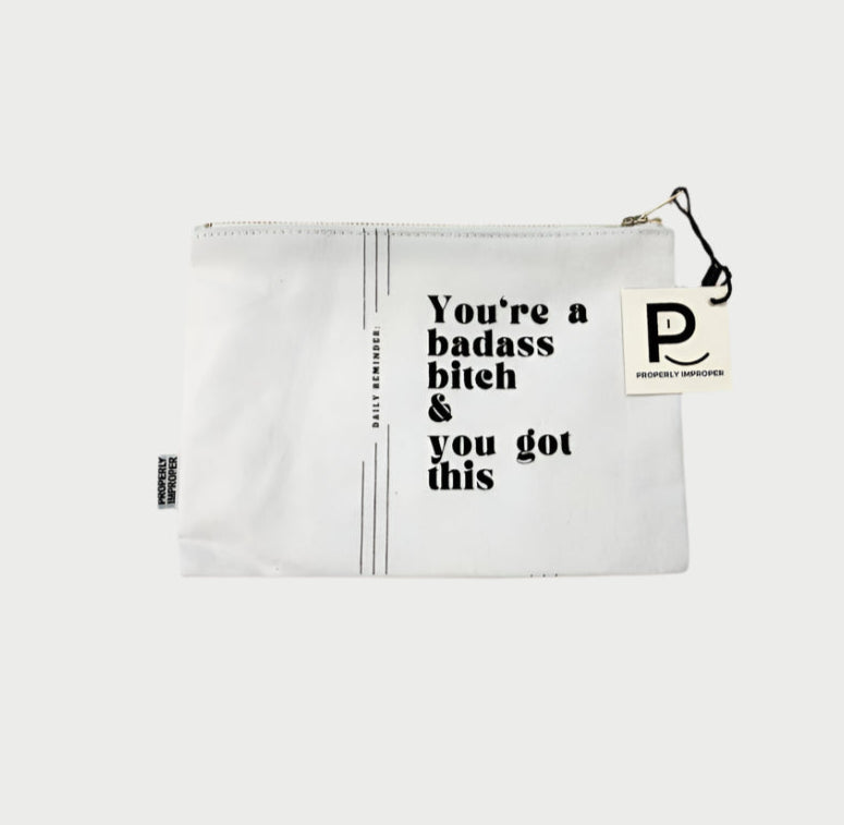 The Improper Pouches by Properly Improper is a durable white canvas pouch featuring a black zipper and tag, ideal as a gift. It boldly displays "You're a badass bitch & you got this" in striking black letters.