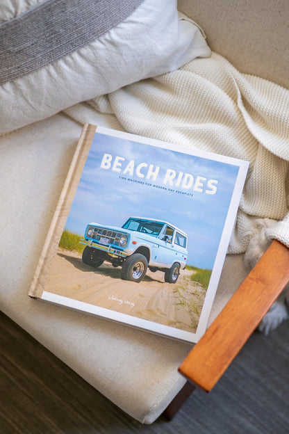 The "Beach Rides" book by Johnny Vacay, depicting island history with a vintage blue SUV on a sandy beach, is placed on a beige upholstered chair with wooden armrests. The chair is complemented by a gray and white pillow.