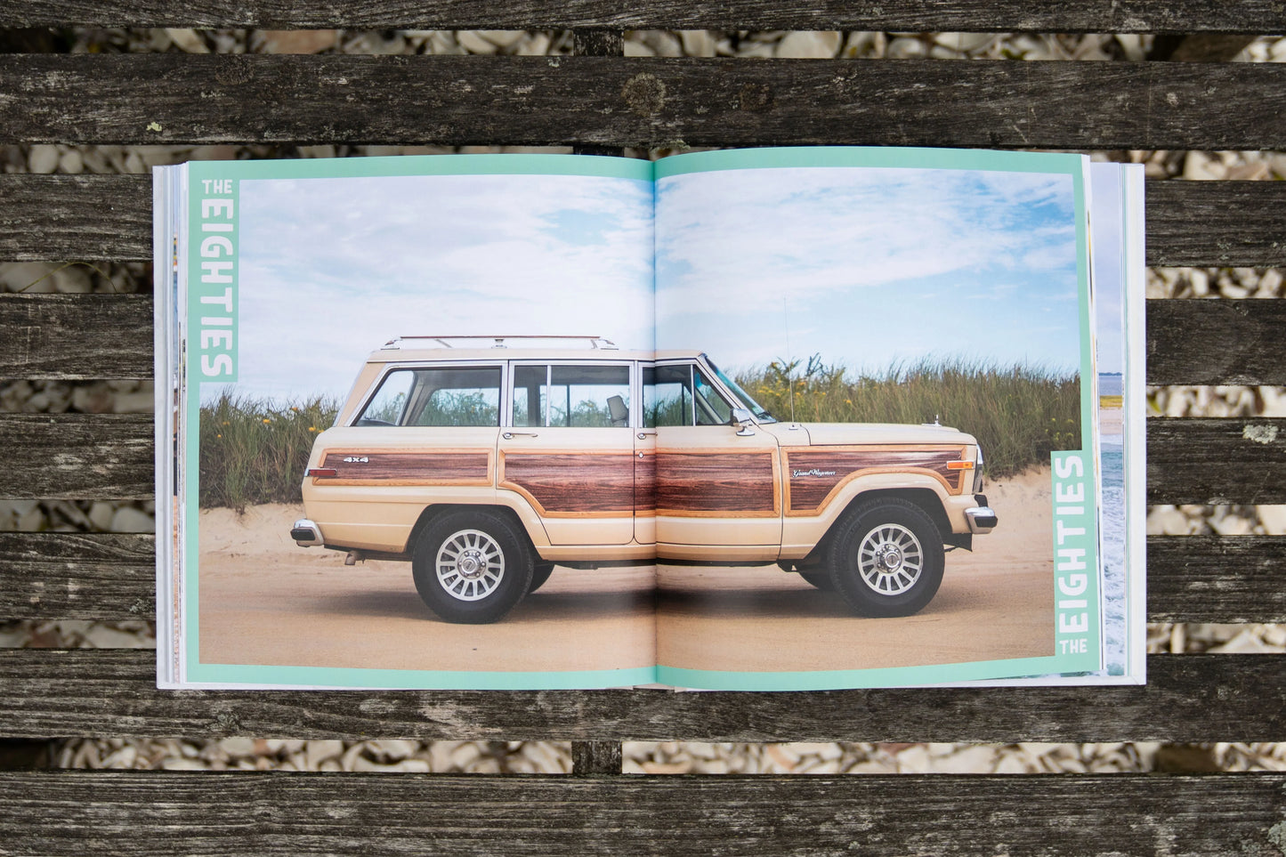 A copy of the "Beach Rides Book" from Johnny Vacay lies open on a wooden surface, showcasing a photograph of a vintage SUV with wood paneling parked on a sandy beach. The scene is complete with tall grass and a blue sky filled with clouds while the title "THE EIGHTIES" stands out, celebrating an era beloved by vintage car enthusiasts.