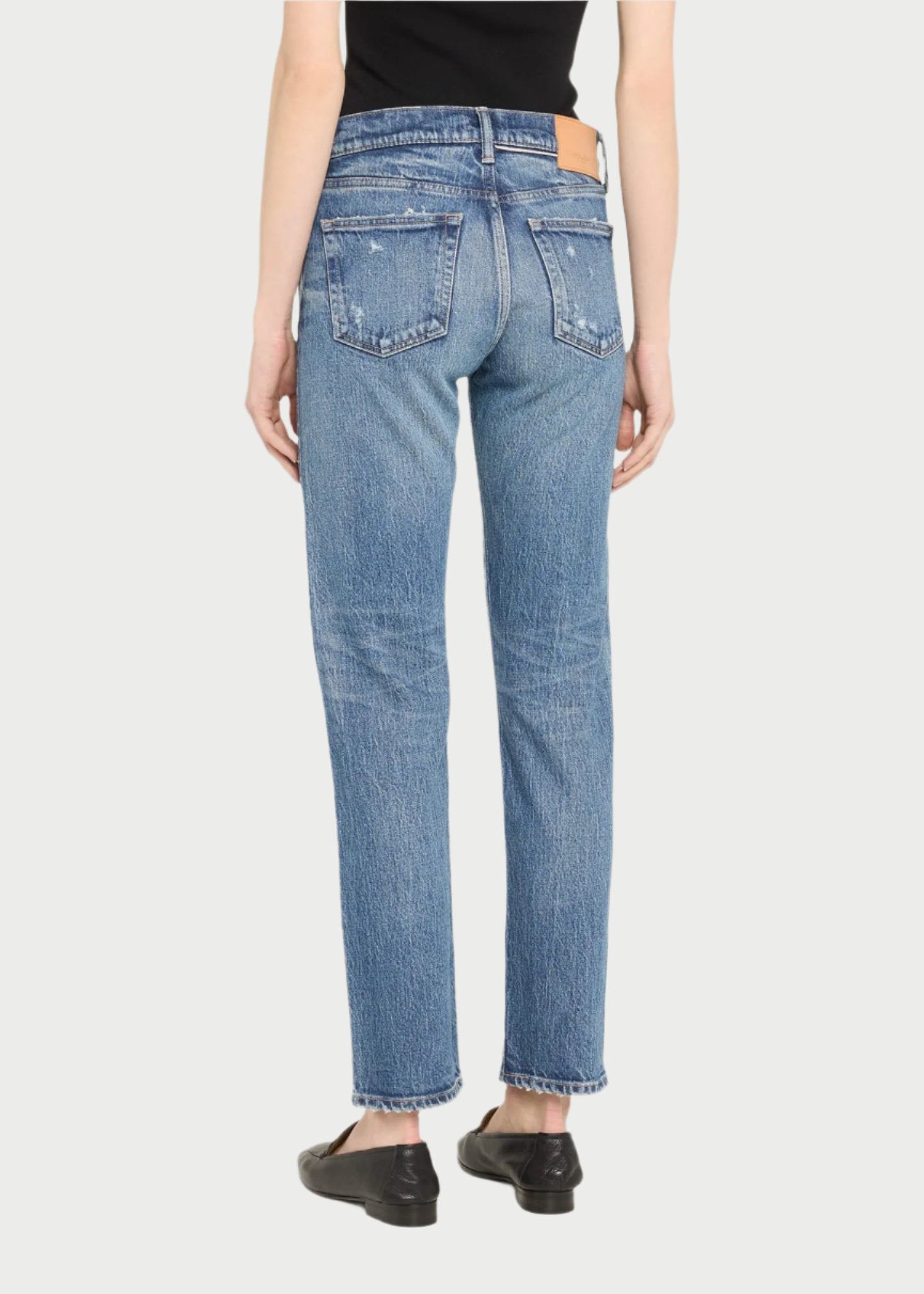 Back view of a person in Moussy's Mallard Straight Jeans, showcasing a medium-wash with a slightly distressed appearance. The jeans feature five pockets and a leather patch on the waistband. They are paired with black flats and a sleek black top.