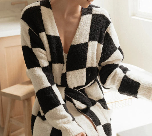 A person sits on a wooden stool, enveloped in the LOCAL BEACH Luxe Robe, which features a checkered black and white Feather Knit design with a waist tie. The softly lit room in the background is illuminated by natural light streaming through the window.