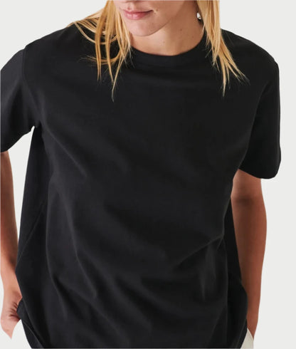 A person with long blonde hair is wearing a Pima Boyfriend Crew T-Shirt in Black by PATRICK ASSARAF. They are standing against a light background, looking slightly downward with their hands in their pockets, highlighting the effortless elegance of this elevated T-shirt crafted from Peruvian pima stretch jersey.