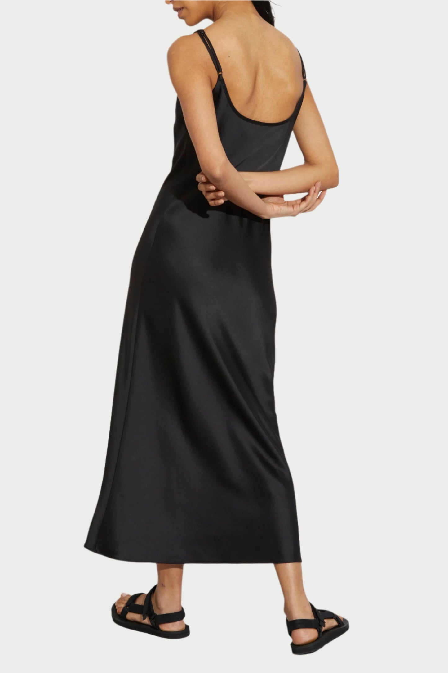 A woman with wet, slicked-back hair poses with her back to the camera, wearing the black, sleeveless Black Haley Slip Dress from Saint Art NY. Her hands are intertwined behind her back and she is wearing black sandals. She stands against a plain white wall.