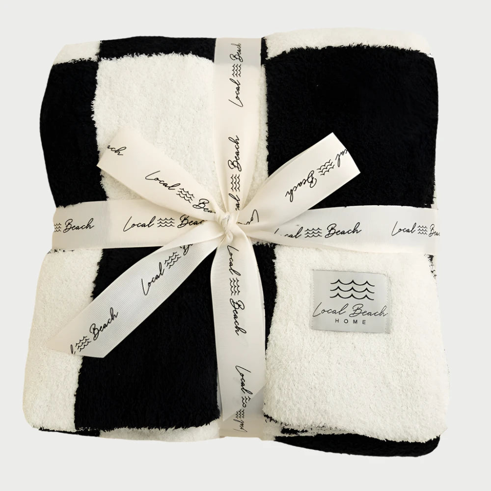 A Luxe Checkered Blanket in black and white, made from soft polyester microfiber, is neatly folded and tied with a ribbon that says "LOCAL BEACH." The XL blanket's label also reads "Local Beach Home.