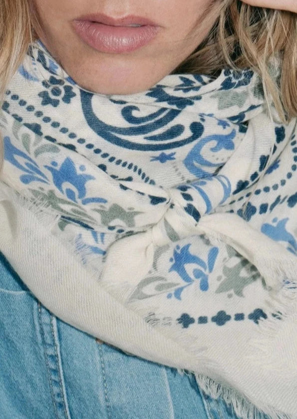 A person wearing round sunglasses and a Maison Lecomte Flament Wool Cashmere Bandana Scarf with a blue pattern. The person has light blonde hair and is dressed in a light denim top. They are looking directly at the camera with a neutral expression, adjusting their sunglasses with one hand.