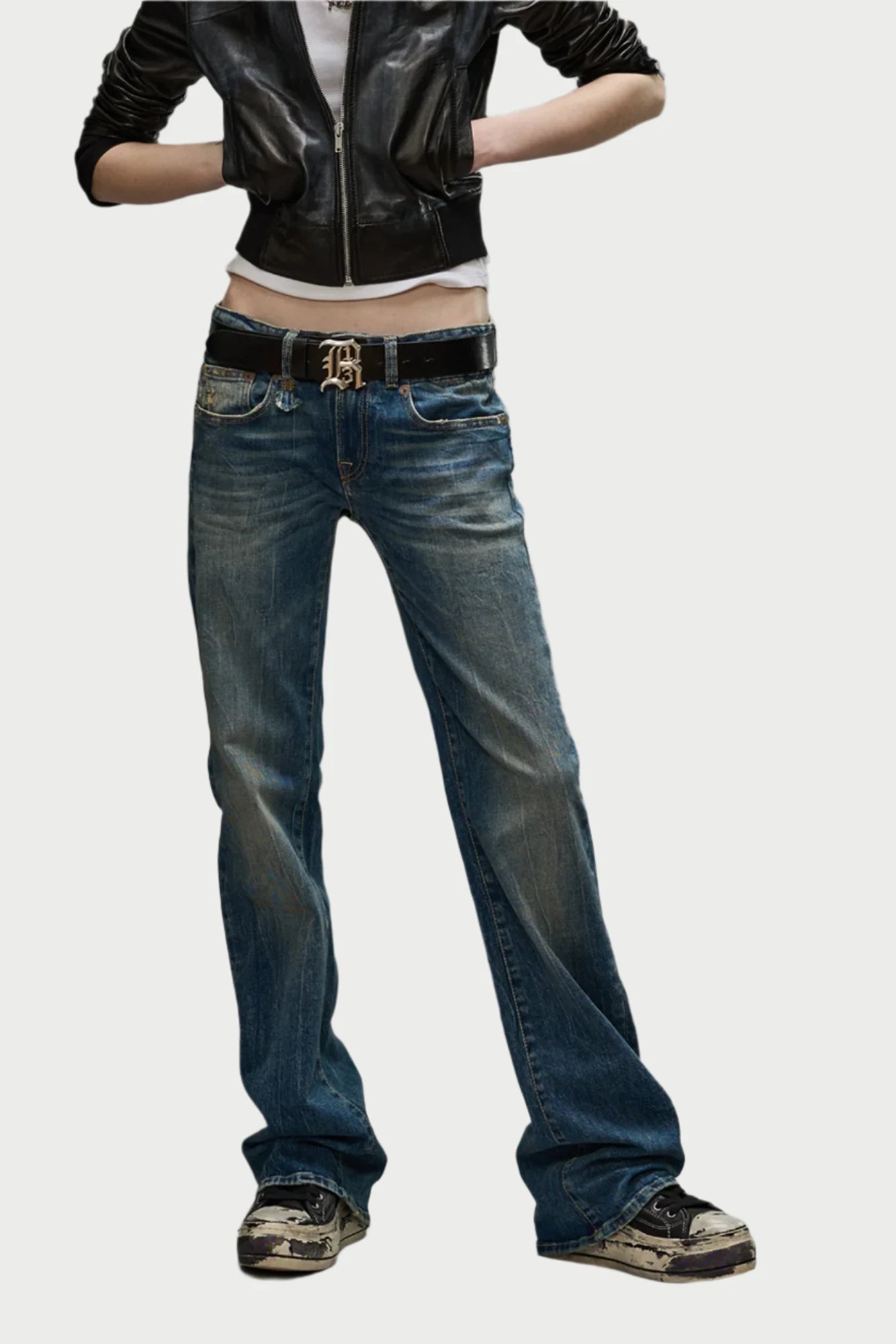 A person confidently stands with arms crossed, dressed in sleek black leather jacket and a stylish white crop top. They wear blue low-rise Boy Flare jeans by R13 paired with a black belt featuring a decorative buckle, and complete the look with trendy black and white sneakers, effortlessly merging style with ultimate comfort against a white background.