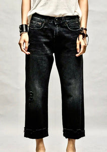 A person wearing black, slightly distressed jeans with a white top. The R13 Jake Black Boyfriend Jean features a relaxed fit with frayed hems and small tears near the knee. The individual has bracelets on each wrist, including a black one on the left and a bundle on the right. The background is plain gray.
