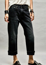 Load image into Gallery viewer, A person is wearing black, slightly distressed R13 Jake Black Boyfriend Jean with frayed hems, a white t-shirt, and black sandals. They have black bracelets on their wrists. The image focuses on the relaxed fit of the premium denim against a plain background.