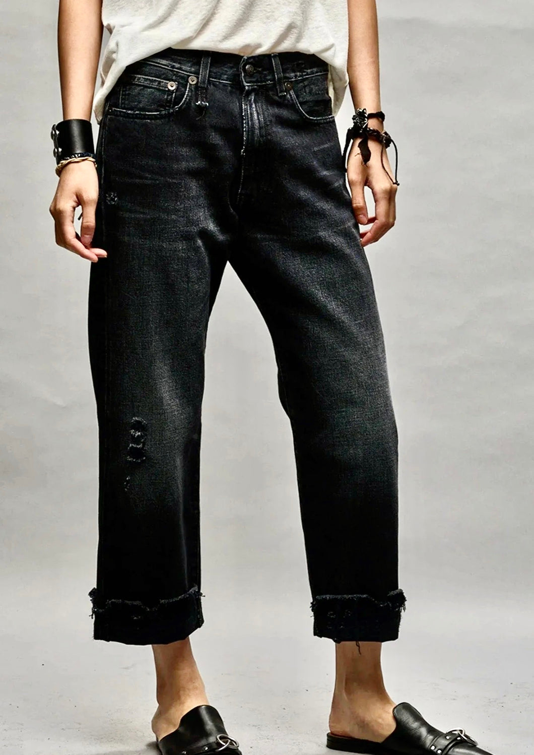 A person is wearing black, slightly distressed R13 Jake Black Boyfriend Jean with frayed hems, a white t-shirt, and black sandals. They have black bracelets on their wrists. The image focuses on the relaxed fit of the premium denim against a plain background.