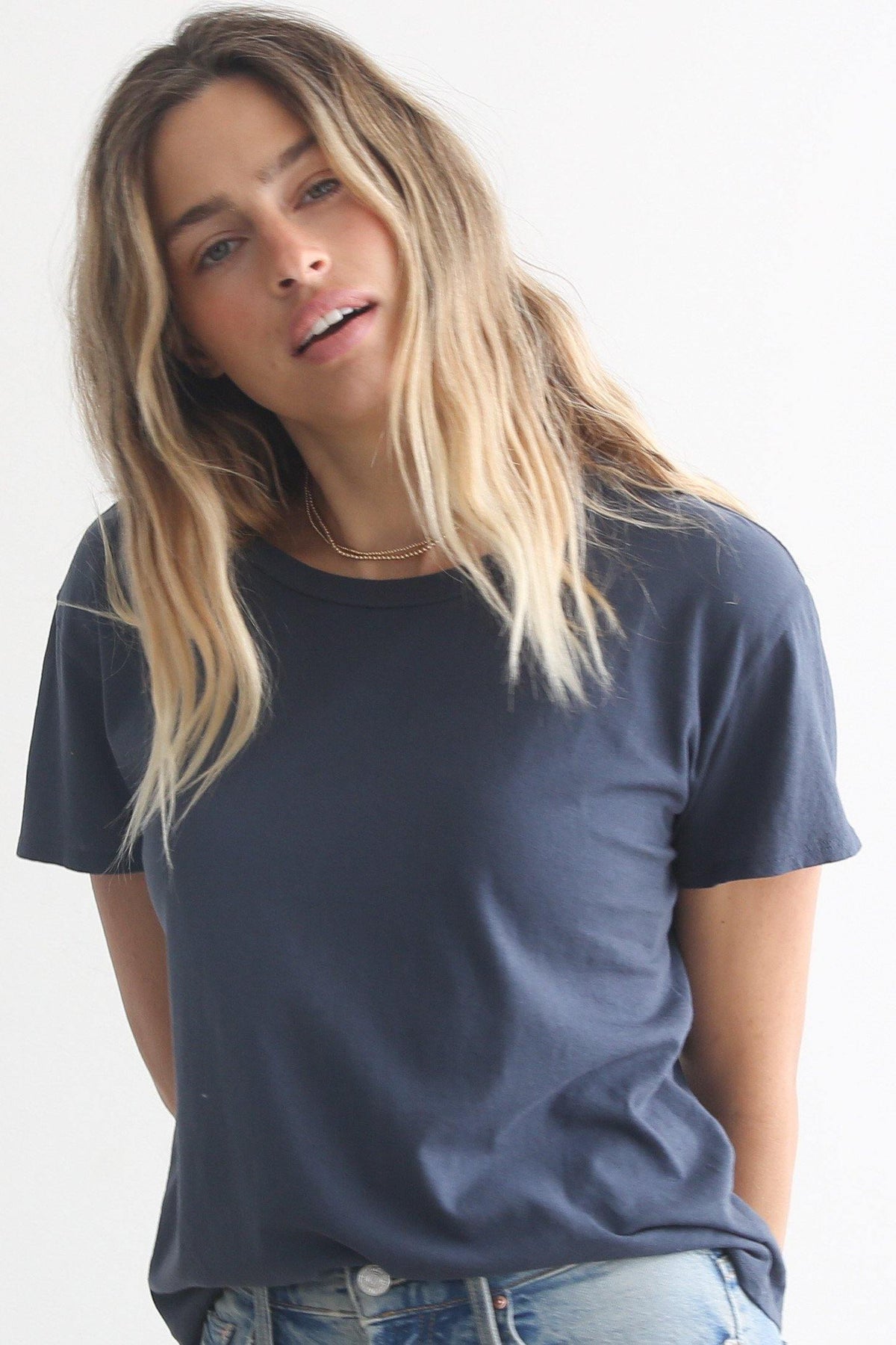 A person with wavy, shoulder-length blonde hair is standing against a plain background. They are casually dressed in a Harley Boxy Crew from perfectwhitetee, layered under a dark T-shirt and light blue jeans, looking directly at the camera with a slightly tilted head.