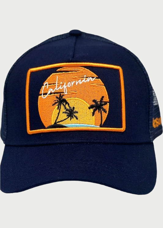 The California Sun Trucker Hat by Soulbyrd boasts a stylish navy blue design with a patch showcasing a sunset, two palm trees, and the word "California" in cursive against an orange and yellow gradient background. This hat features adjustable snap back closures and black mesh back panels for added comfort.