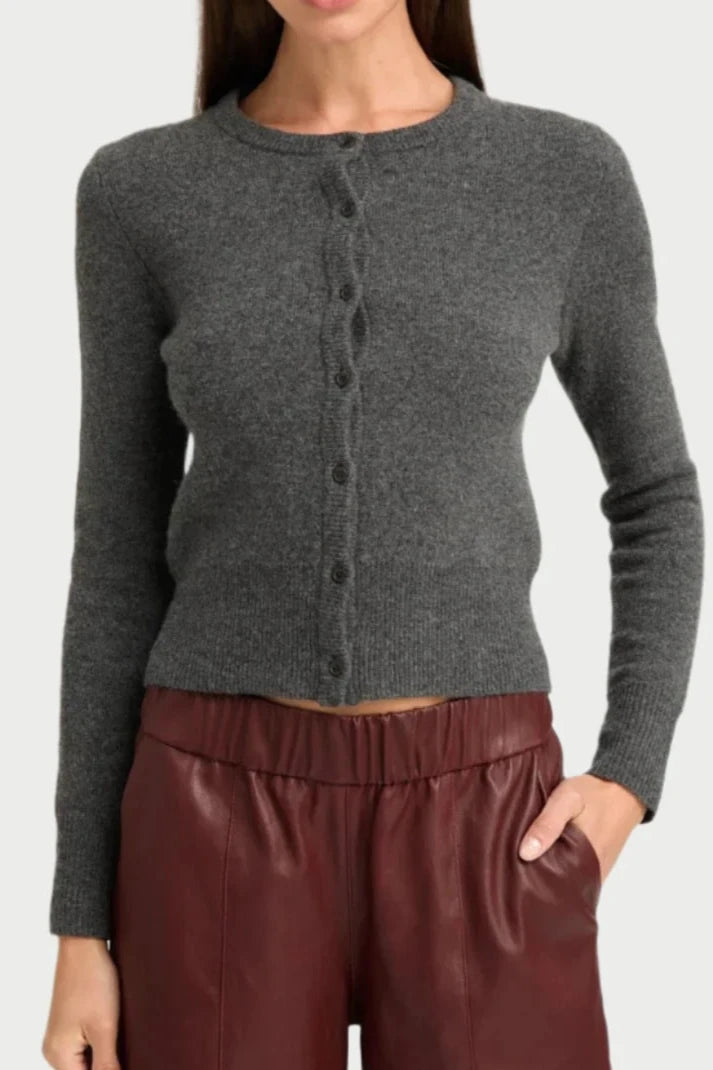 The person wears a charcoal baby cardigan and maroon leather pants from SPRWMN. With one hand tucked into a pocket, the ultra-soft ensemble stands out against the plain backdrop.