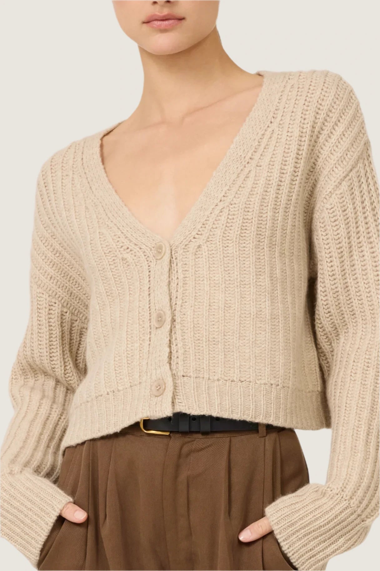 A person showcases casual elegance, stylishly dressed in the Polly Cashmere Cardigan by One Grey Day. The cardigan features a deep V-neck and buttons, perfectly paired with high-waisted brown pants against a plain light-colored backdrop.