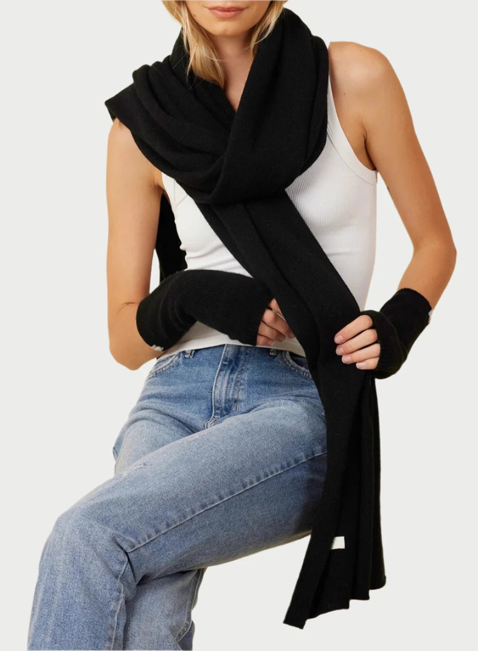 A person with wavy blonde hair, wearing a white tank top and blue jeans, sits on a wooden stool. They have a luxurious Cashmere Travel Scarf from One Grey Day draped around their neck and black fingerless gloves on both hands. The background is a plain light beige color.