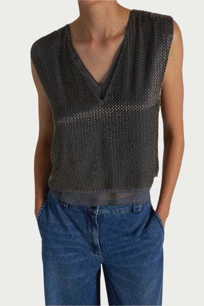 Person wearing a sleeveless, semi-transparent, textured gray Cassidy Beaded Top by Saint Art NY over a V-neck inner shirt, paired with blue jeans. The person has their hands in their pants pockets. The background is plain and light-colored.