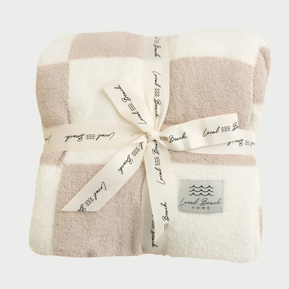 A folded Luxe Checkered Blanket in beige and white, which is reversible, is tied with a printed ribbon. The label reads "LOCAL BEACH." This XL blanket is Oeko-Tex certified, guaranteeing high safety standards for your comfort.