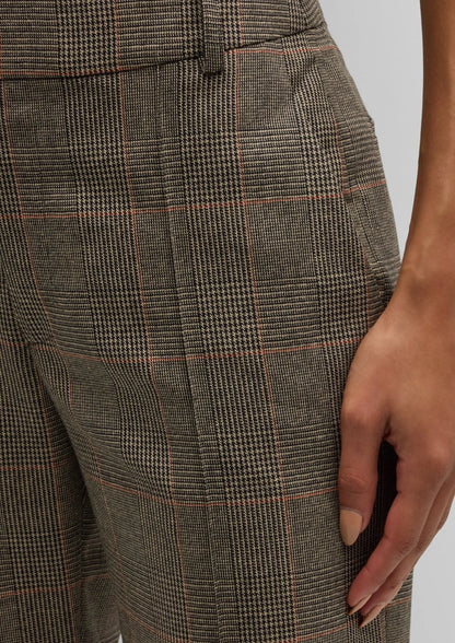 Close-up image of a person wearing FRAME's Check Ankle Trouser, featuring high-waisted, flared-leg plaid trousers with a classic tartan pattern in shades of brown and grey, accented by subtle red lines. The person’s right hand rests on the thigh, drawing attention to the side pocket of the stylish garment.