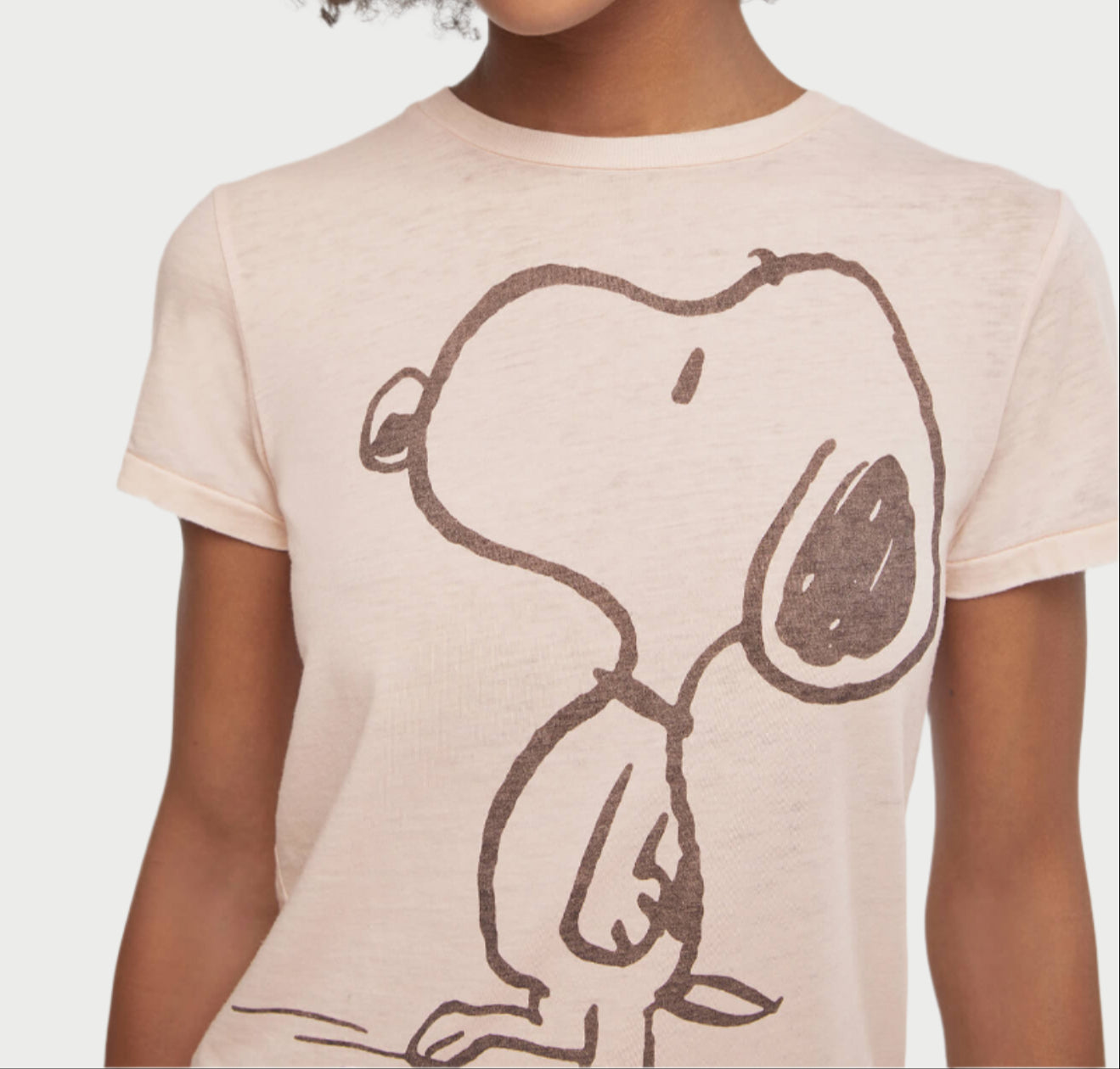 Wearing the Dahlia Snoopy Tee by RE/DONE, a casual light pink cotton-polyester blend shirt featuring a bold outline of the cartoon dog Snoopy on the front, only the upper torso is visible against a plain white background.