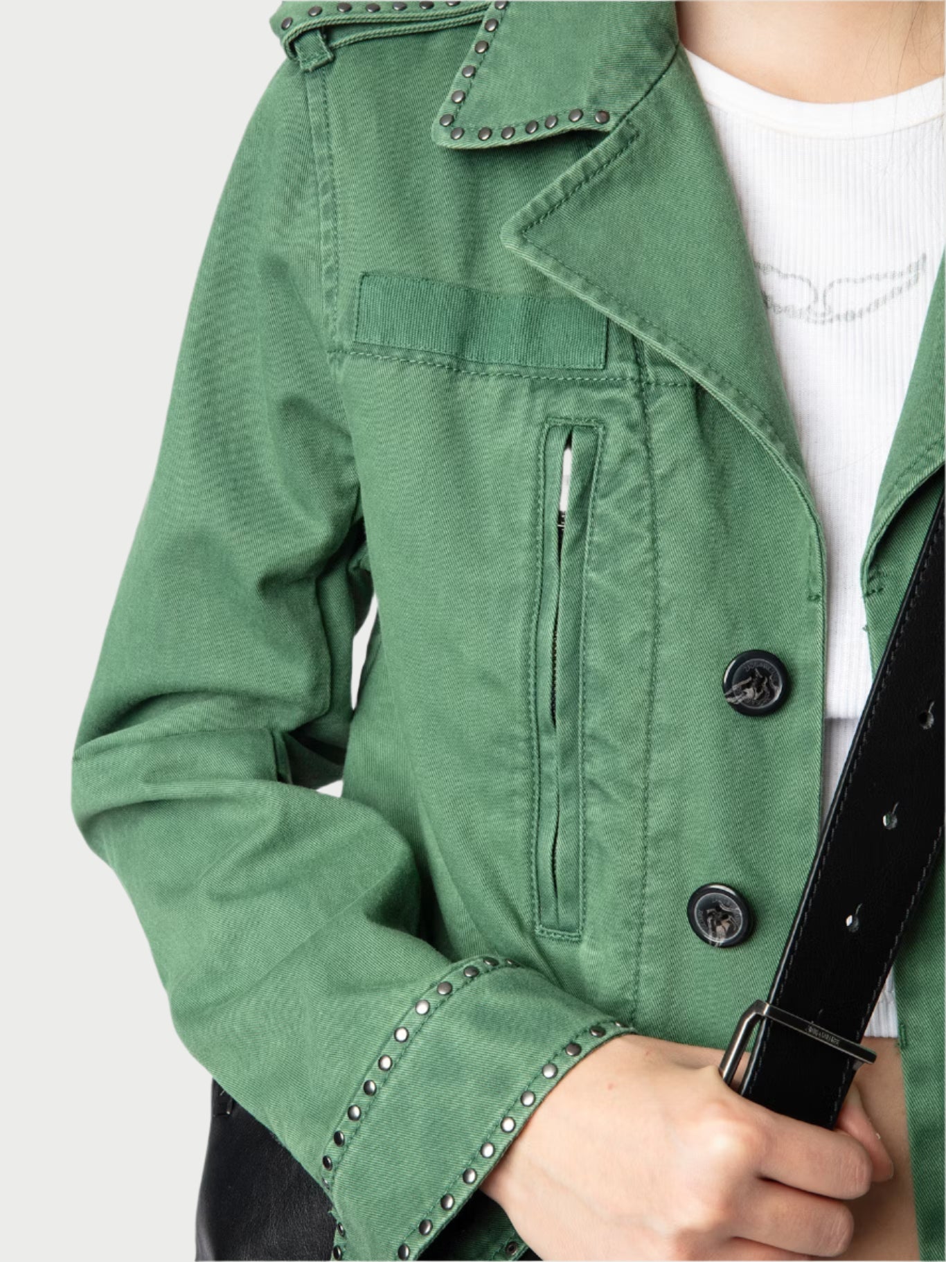 A person wears an army green Klimi Utility Jacket by Zadig & Voltaire with metal studs on the shoulders and collar. The buttoned jacket complements a white shirt underneath, while the person holds a black strap, likely from a bag.