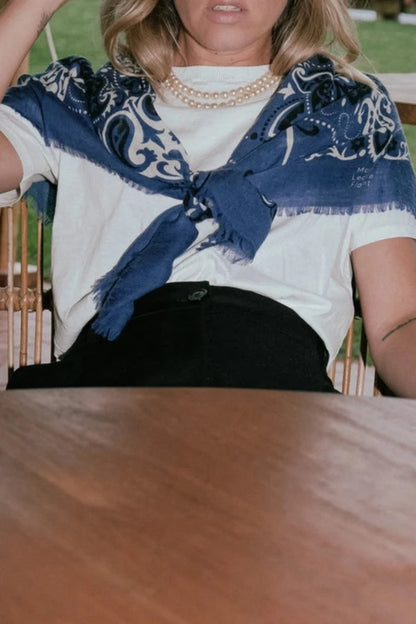 A person with shoulder-length blond hair sits at an outdoor wooden table wearing a white shirt with a Maison Lecomte Flament Wool Cashmere Bandana Scarf, black sunglasses, and a pearl necklace. The background includes a grassy area with blooming flowers and a distant view of water.