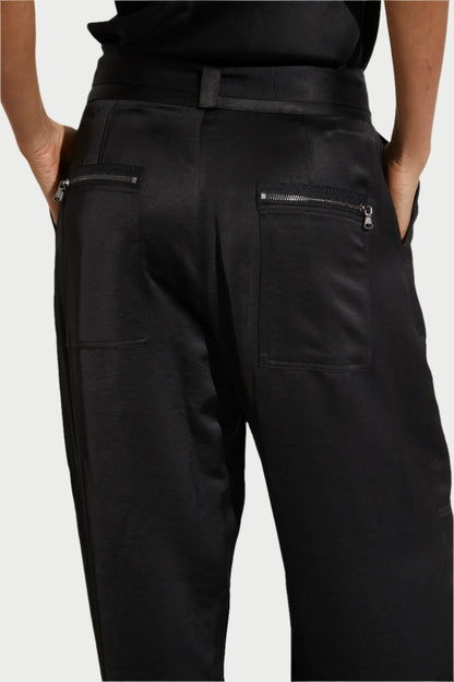 The image features a person wearing Saint Art NY's Charmeuse Pants, highlighting the sleek and slightly shiny black fabric. These pants come with utility pockets, including two rear zipper pockets. The outfit is completed with a tucked-in black shirt, set against a neutral background.