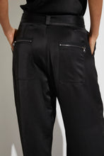 Load image into Gallery viewer, A person is wearing the Charmeuse Pant by Saint Art NY, featuring a sleek black design with cargo-inspired back pockets that have zipper closures. Their upper body is partially hidden, and their hands rest on their hips beside the pants. The shiny, smooth texture of the Charmeuse Pant adds an elegant touch to the overall look.