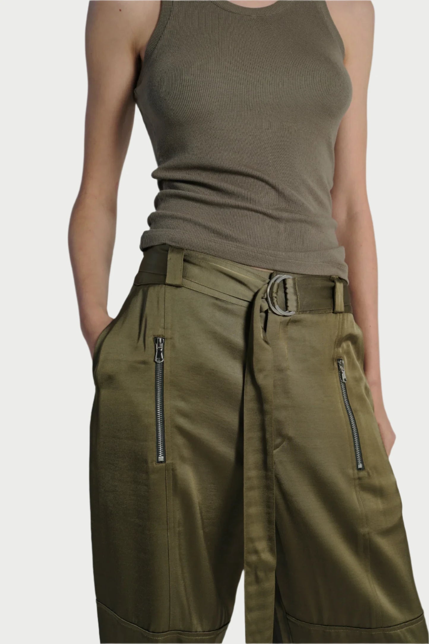 A person wearing a gray sleeveless top and olive green Saint Art NY Charmeuse Pants, featuring utility pockets and a detachable d-ring belt, with their hands casually tucked in the pockets.