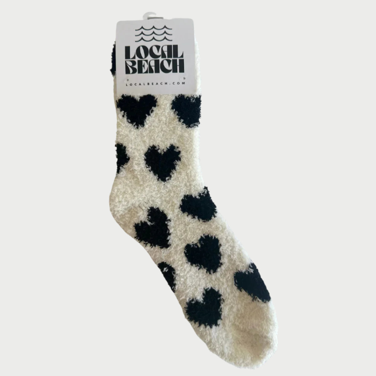 The "LOVE Socks" by LOCAL BEACH are perfect for Galentine's Day, featuring black heart patterns on a cream background. Each fuzzy sock comes with a cardboard tag reading "Local Beach.