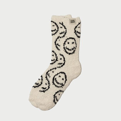 The Cozy Socks by LOCAL BEACH is a fluffy cream slipper sock with a black smiling face pattern.