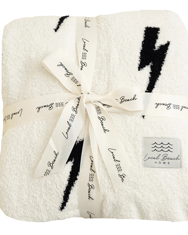 The Luxe Lightning Bolt Blanket by LOCAL BEACH is a soft, white reversible XL blanket featuring black lightning bolt patterns. It's neatly folded and secured with a ribbon printed with "LOCAL BEACH" in black. Crafted from 100% polyester microfiber, this blanket is OEKO-TEX Standard 100 certified. A label in the bottom corner reads "LOCAL BEACH home," accompanied by a wave design.