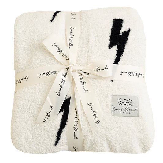 The Luxe Lightning Bolt Blanket by LOCAL BEACH is a soft, white reversible XL blanket featuring black lightning bolt patterns. It's neatly folded and secured with a ribbon printed with "LOCAL BEACH" in black. Crafted from 100% polyester microfiber, this blanket is OEKO-TEX Standard 100 certified. A label in the bottom corner reads "LOCAL BEACH home," accompanied by a wave design.