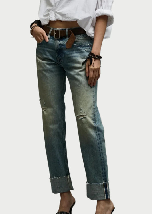 A person is wearing R13's classic slim fit, faded, Cuffed Romeo Jean with a distressed look and a white, cropped blouse. They are accessorized with a brown belt, a black bracelet, and black high heels. The background is plain and gray.