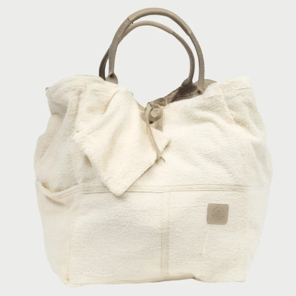 A large, beige Reverse Terry Bag made by Travaux en Cours is made from soft, textured cotton canvas. It features sturdy handles, an open top secured by a small strap, side pockets, and a stitched patch near the base with a logo. The bag is slightly slouched, perfect for beach days with its casual, relaxed appearance.