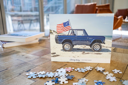 The Johnny Vacay 500 Piece Puzzles box displays an image of a blue off-road vehicle adorned with an American flag, parked on a beach. Puzzle pieces are scattered in front of the box on a wooden table, evoking the spirit of family fun that Johnny Vacay is known for, with furniture and a stack of books blurred in the background.