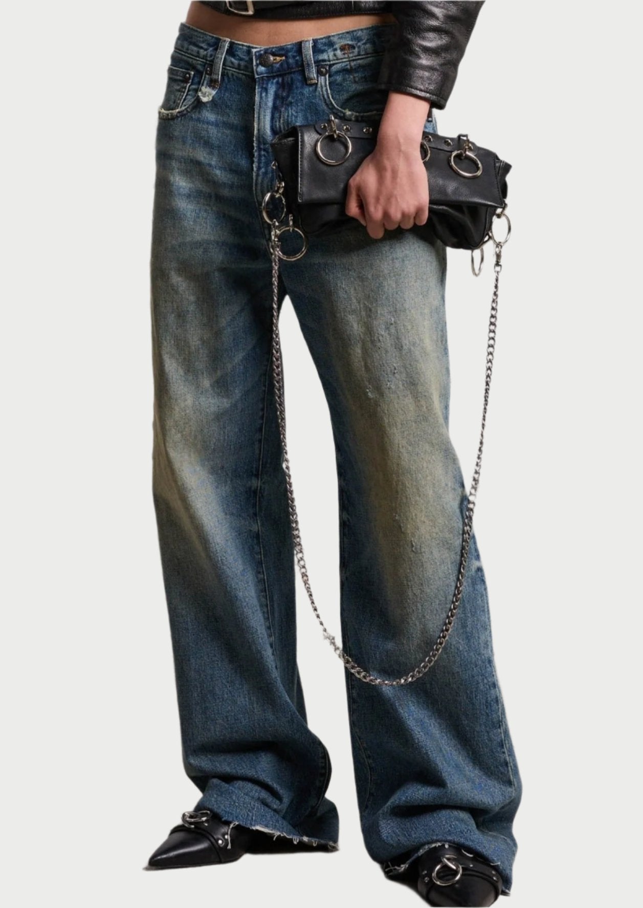 Person wearing distressed wide-leg D'Arcy Loose Jean by R13 in Japanese Indigo Denim, a black leather jacket, and pointed black shoes with metal details, holding a black clutch bag with silver chains and ring embellishments. The background features a white brick wall with some wear.