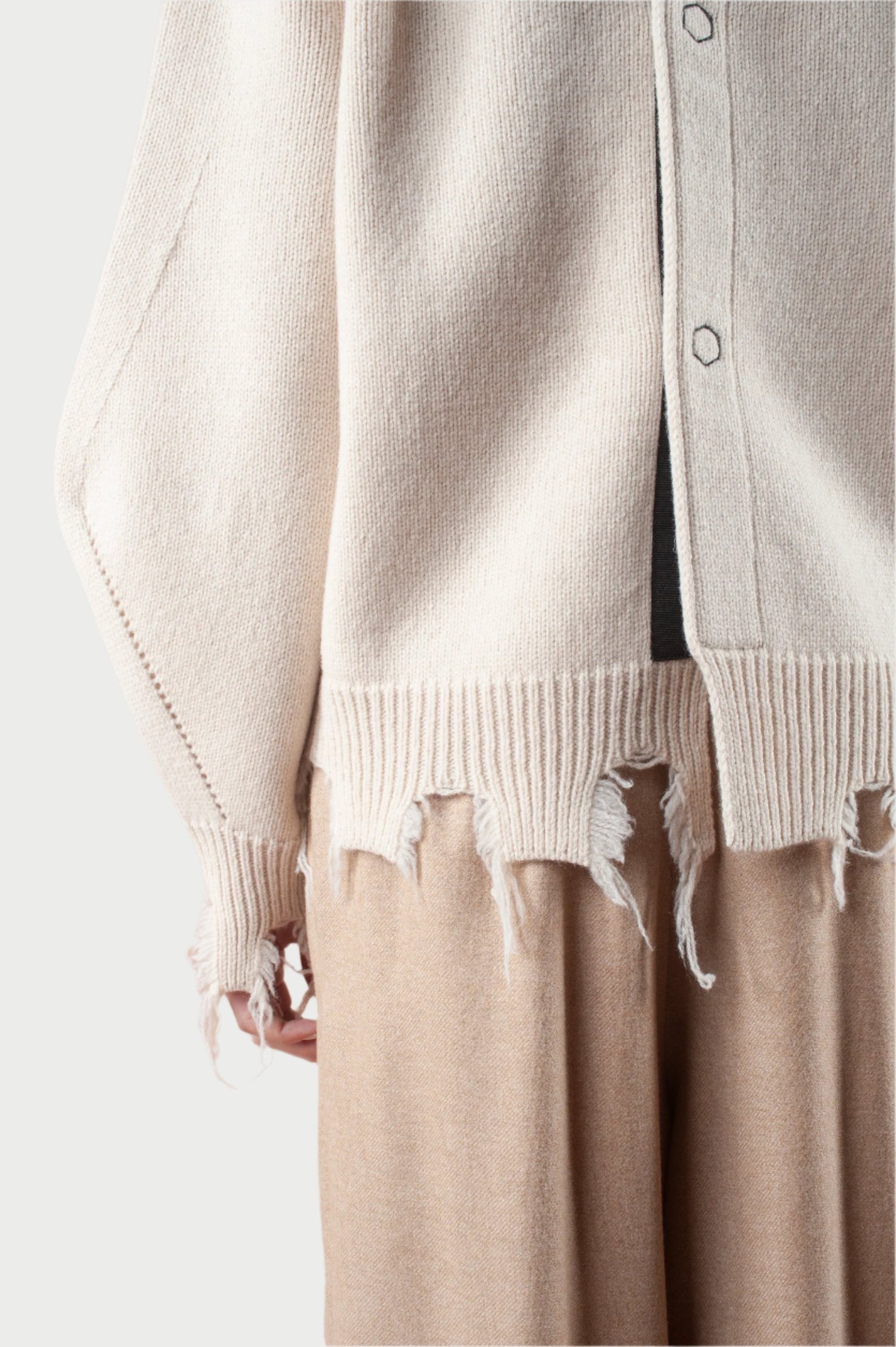 A person wearing the Turnable Sweater w/Banana Sleeves by Phisique du Role, characterized by its beige knit material with frayed hems and loose threads, paired with wide-legged tan pants. The sweater features buttons and ribbed cuffs. The image emphasizes the texture and intricate details of the clothing.